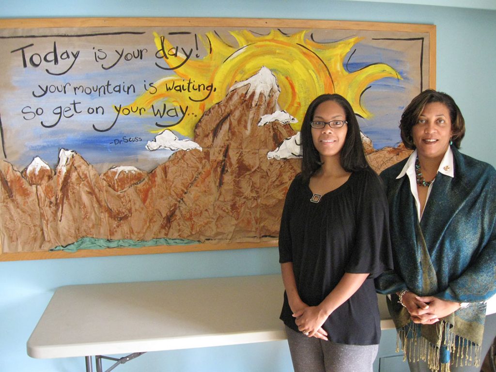 Tasha Melvin (left) and Dianne Pledger are both employed by Genesis Home, a transitional housing facility in Durham for those experiencing homelessness. Here, Melvin and Pledger stand by a painting in the home’s conference room. It contains a quote from Dr. Seuss: “Today is your day! Your mountain is waiting, so get on your way.” It’s a constant reminder to both the employees and the residents at Genesis Home that each day is an opportunity to better oneself.